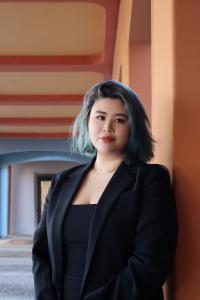 Headshot of Xinyi Tong, the 2025 Outstanding Communication Senior. Tong's hair is should length and fades from teal to black. The are wearing a black blazer, and standing outside the entrance to the Main Theatre at Wright Hall