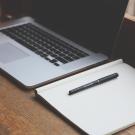 Wood desk, wtih Mac laptop, and a notebook and pen. 
